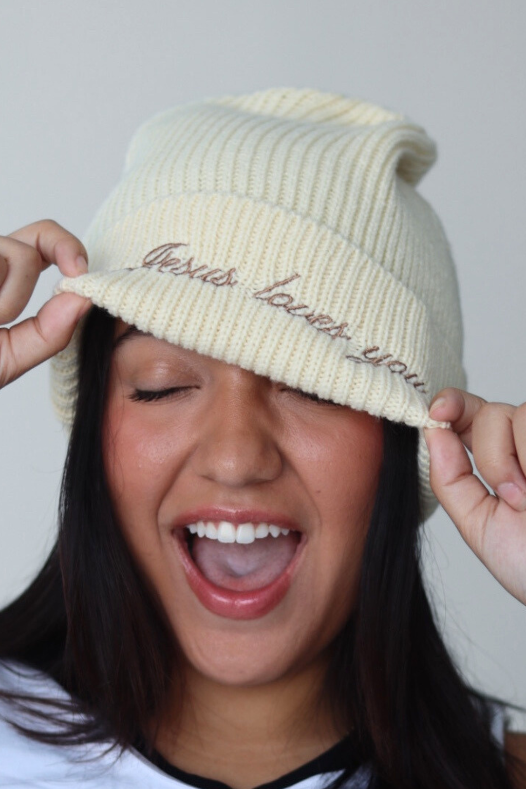 Person wearing a cream-colored knit beanie with text, smiling against a plain background