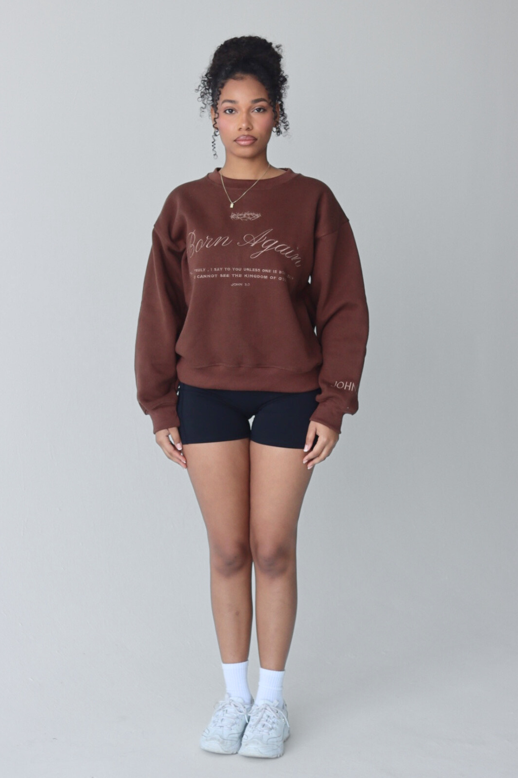Woman wearing a brown sweatshirt with text, standing against a plain background